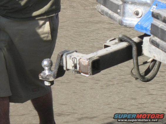 Trailer hitch receiver with ball mount and safety chain loop. Person in shorts stands nearby