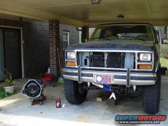 Dark blue Ford Bronco on jack stands, front end disassembled, with a transmission on a jack nearby