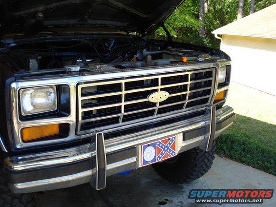 Front grille and bumper of a Ford truck, hood open showing engine components. License plate frame displays a Confederate flag