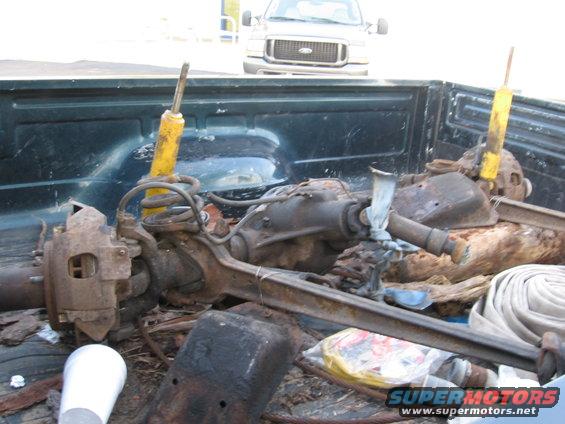 Front axle assembly with yellow shocks and springs in truck bed. Another truck is visible in background