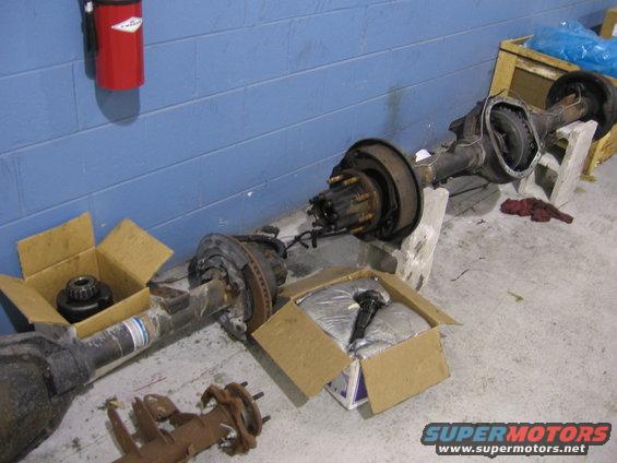 Two vehicle rear axles are on the floor, one in a box. A red fire extinguisher is on the wall