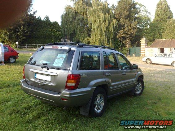 Silver Jeep Grand Cherokee parked on grass, rear quarter view. A red car is visible to the left