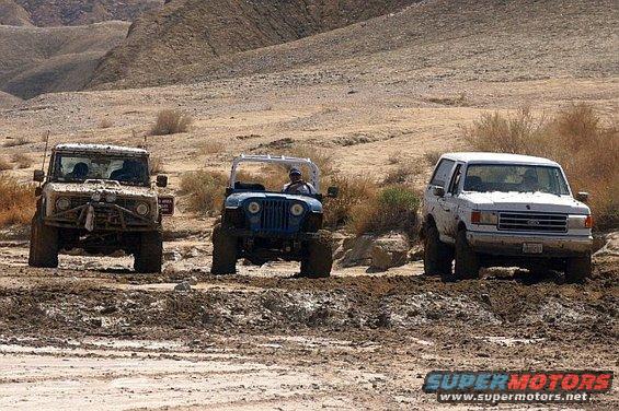 White Ford Bronco, blue Jeep, and a tan off-roader driving through mud