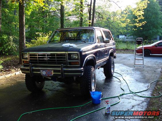Dark blue Ford Bronco with aftermarket wheels and tires, parked in a driveway with a red car in the background