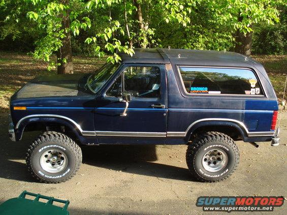 Dark blue Ford Bronco parked outside with trees in the background. Has a 'Bronco' sticker on the rear window