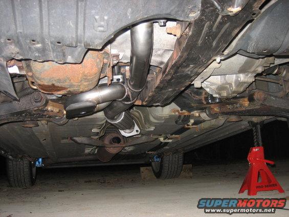 Underside view of a car's exhaust system, showing pipes and mufflers. A red jack stand is visible