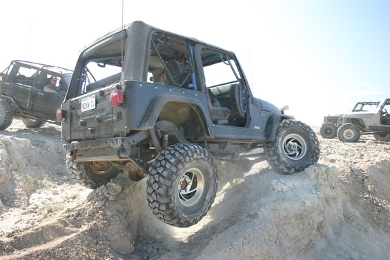 Dark gray Jeep TJ with lifted suspension and large tires climbing a dirt hill. License plate visible