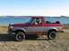1970 Mercury Cougar - A two-door SUV with a red upper body and silver lower body sits parked near a body of water with two dogs visible inside.