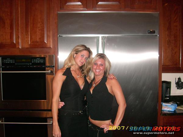 Two women pose in a kitchen in front of a stainless steel refrigerator
