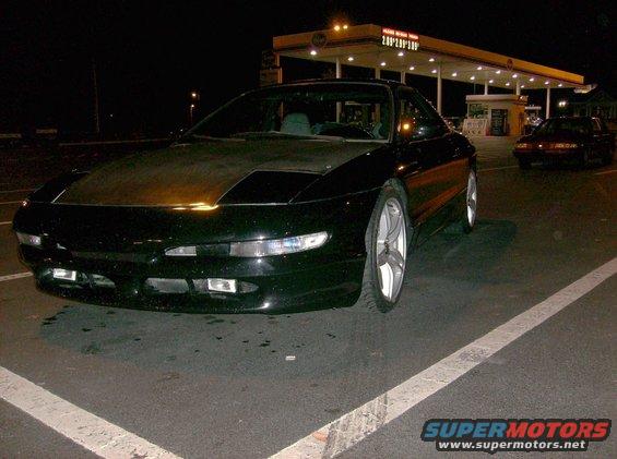 Black coupe with a matte hood parked at a gas station at night. Another car is visible in the background