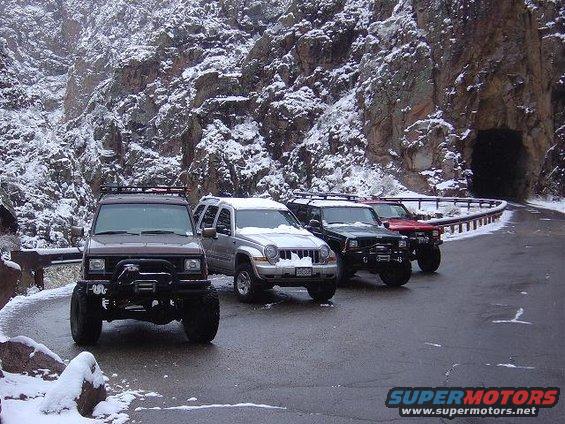 Four SUVs parked on a snowy mountain road next to a tunnel entrance. The lead vehicle is dark brown, followed by silver, black, and red SUVs