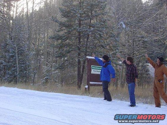 Three men point towards a sign in a snow-covered forest