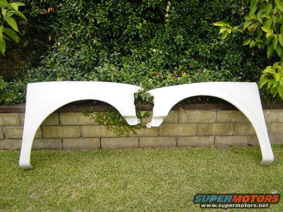 Two white car fenders laid on grass in front of a brick wall and hedge