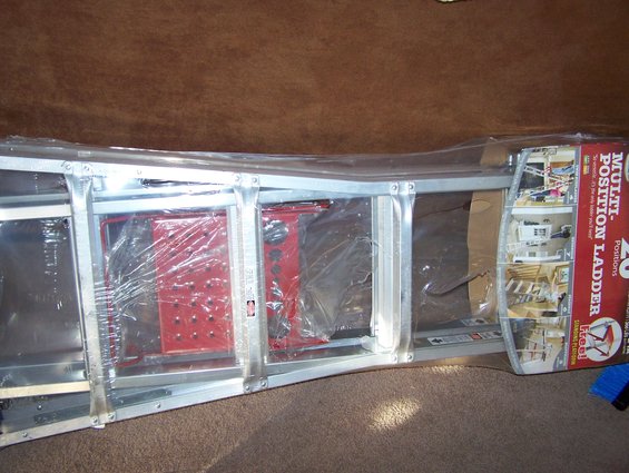 Multi-position ladder in plastic packaging, showing red locking mechanisms and aluminum frame. Box has product images and text