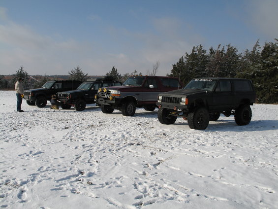 Four modified SUVs parked in a snowy field. The closest is a black Jeep Cherokee. Behind it is a maroon Ford Bronco, then two black SUVs
