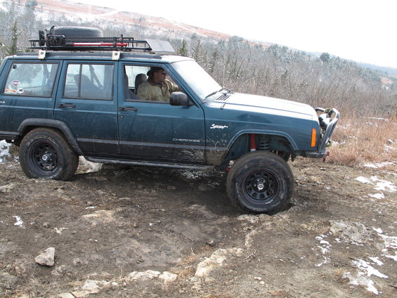Teal Jeep Cherokee Sport with roof rack and spare tire, driving on a dirt trail