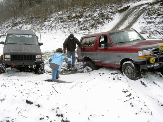 Red and gray Ford Bronco stuck in snow, front right tire deep in mud, with a black Jeep Cherokee nearby