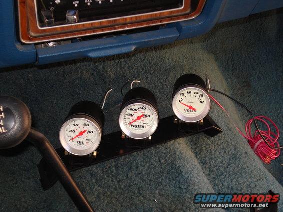 Three aftermarket gauges (oil, water, volts) mounted on a black bracket in a vehicle's interior, next to a gear shift