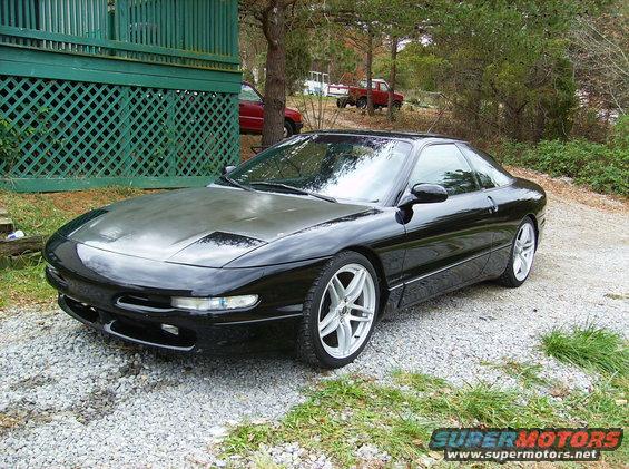 Black 1995 Ford Probe parked on gravel in front of a green lattice-covered structure. Two red trucks are visible in the background