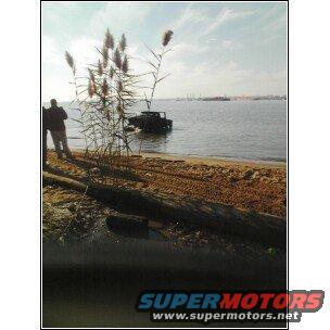 Vehicle partially in water on a beach with two people standing nearby. Tall grass in foreground