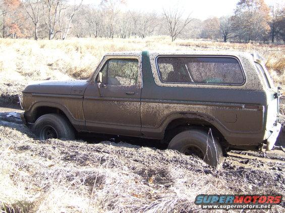Muddy 1978 Ford Bronco stuck in a ditch with dry grass and trees in background