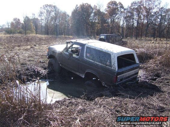 Mud-covered dark gray and white Ford Bronco stuck in a muddy ditch with a black Jeep in the background