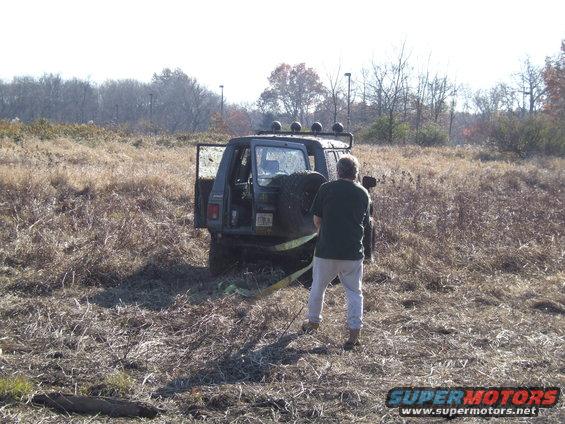 guy trying to pull out a dark SUV stuck in tall dry grass, spare tire mounted on back
