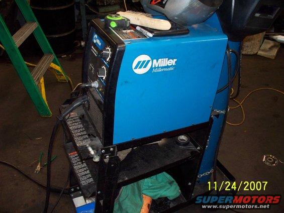 Blue Miller Millermatic welding machine with controls and cables, on a black metal stand. Date stamp 11/24/2007