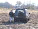1979 Ford Bronco - A person opens the door of a dark green SUV stuck in a muddy, grassy field. The vehicle has a spare tire mounted on the rear.