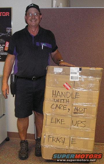Man in FedEx uniform holding stacked boxes with handwritten text 'HANDLE WITH CARE, NOT LIKE UPS FRAGILE'
