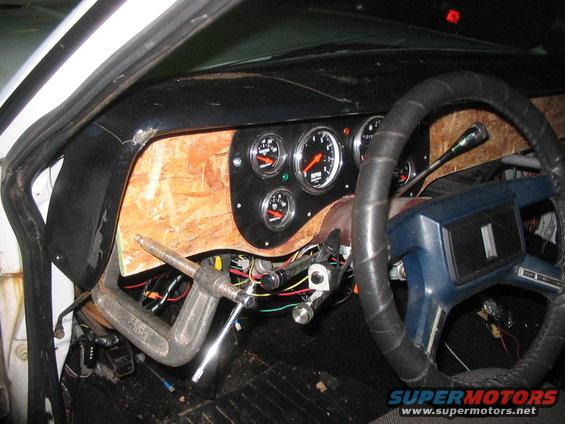 Custom dashboard with aftermarket gauges and plywood paneling. Steering wheel is wrapped in black tape