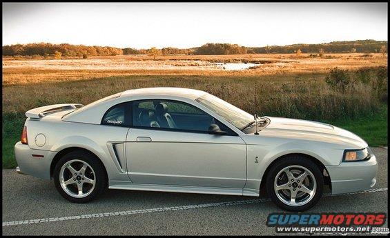 Silver Ford Mustang parked on a road with a field and water in the background