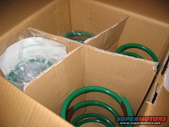 Four green coil springs in a cardboard box with dividers. Some are in plastic bags