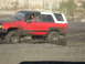 1993 Ford Bronco - A red and black SUV navigates through a muddy terrain, kicking up water and dirt.