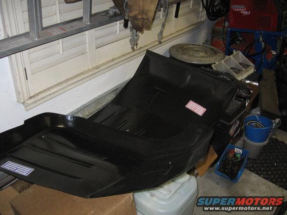 Black metal floor pan with two white labels, resting on boxes in a garage
