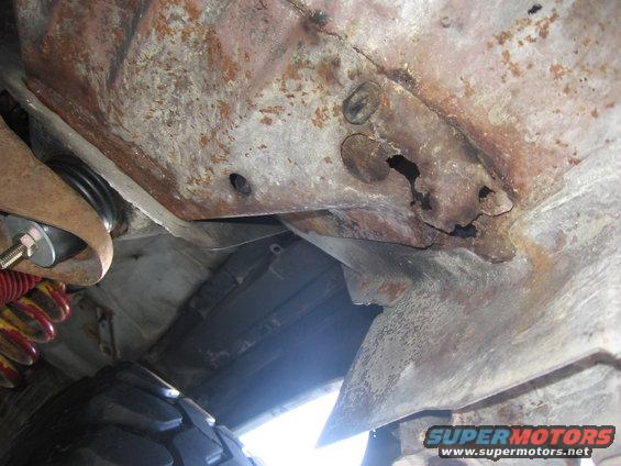 Heavily rusted metal frame with holes, part of a vehicle undercarriage. A tire is visible