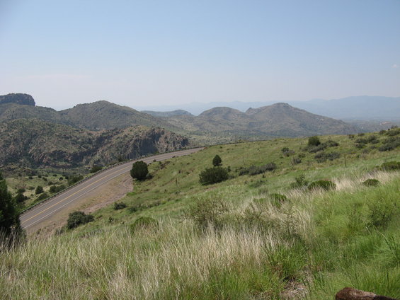 Winding asphalt road through a grassy, hilly landscape with mountains in the background under a clear sky