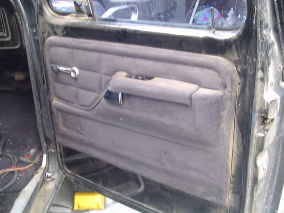 Interior of a dark-colored vehicle's driver side door panel, showing gray fabric upholstery, a chrome window crank, and a black armrest