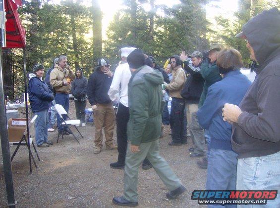 Group of people in casual clothing gathered outdoors in a wooded area, with a red tent on the left