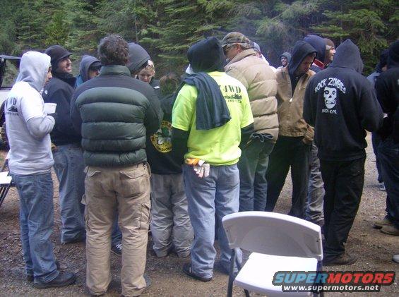 Group of people in casual outdoor clothing, some wearing hoodies, gathered in a wooded area. A white folding chair is in the foreground