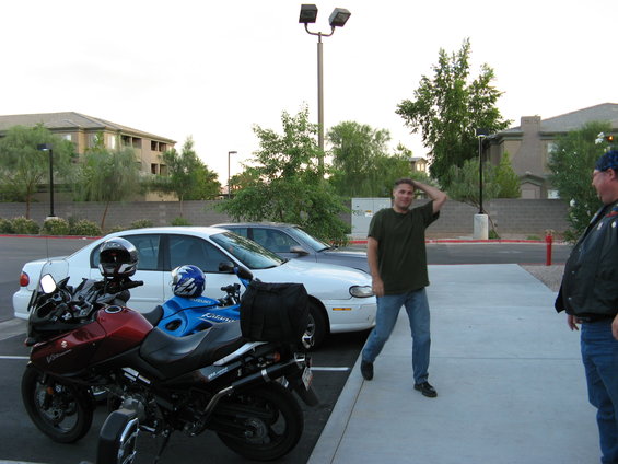 Two motorcycles, one red and one blue, parked next to a white car and a dark car. Two men stand on a concrete walkway