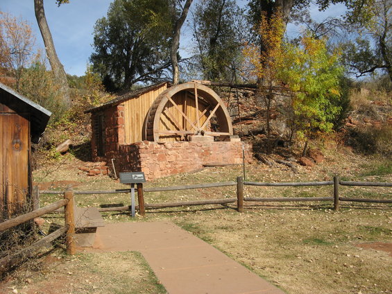 Old stone and wood building with a large wooden water wheel, surrounded by trees and a wooden fence