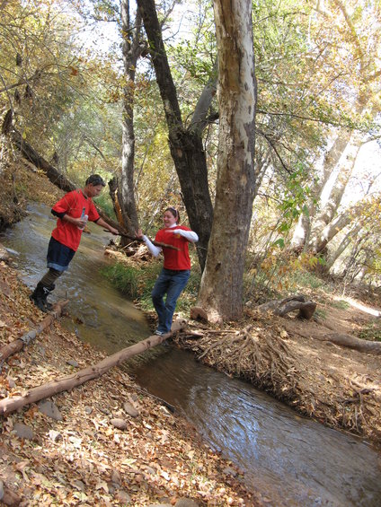 Two people in red shirts crossing a stream on a fallen log, surrounded by trees with autumn leaves