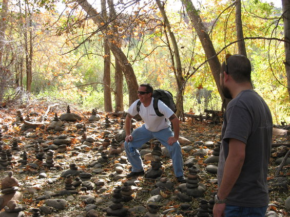 Man in white shirt and blue jeans crouches near stacked rocks in a forest. Another man stands to the right