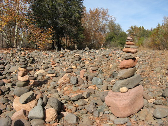 Rock cairns stacked on a rocky, dry riverbed with trees in the background