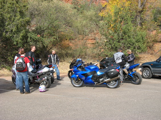 Group of people standing around several motorcycles parked on asphalt, with trees and red rock formations in the background