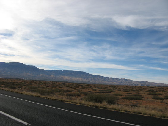 Asphalt road with white lines curves through a dry, brush-covered landscape with distant mountains under a cloudy blue sky
