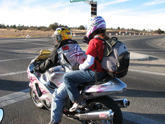 Two people on a silver motorcycle with purple flame graphics. Rider wears a yellow helmet, passenger wears a purple helmet. Aftermarket Yoshimura exhaust visible