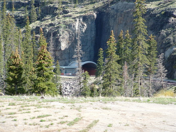 Tunnel entrance with a modern building at its base, surrounded by pine trees and a rocky mountain face