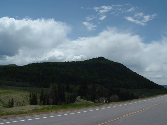 Mountainous terrain with a forest-covered peak and a paved road in the foreground under a cloudy sky